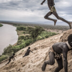 South Ethiopia. 2011. Omo Valley. Along the oriental shore of the Omo River near the Karo village, children are playing by jumping in the sand. The Karo village is located in a natural bights of the Omo River. Karos are a small tribe with an estimated population between 1,000 and 3,000 people and lives thanks to fishing and cultivation made possible by the flooding of the Omo River. Currently, the river forest seen in the background has been demolished and replaced by an extensive cotton plantation of a foreign private company. Following the construction and commissioning of the Gibe III dam, the flooding of the Omo River has stopped, depriving the Karo population of the possibility of cultivating those products that today they’re forced to buy at the market.